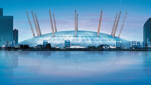 A large stadium with a distinctive roof structure and multiple tall cranes, surrounded by modern buildings and a calm body of water. - The Railway Tavern Hotel (London)