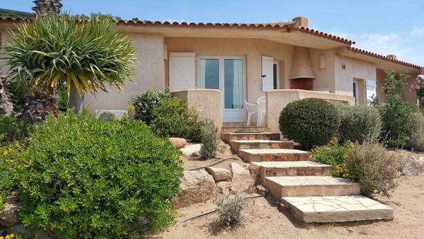 A house with a tiled roof, a front door, and a small porch. There are steps leading to the door and a garden with various plants and shrubs. - Mini-villa No. 9 - 2/4 people (PORTO VECCHIO)