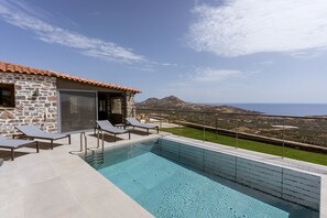 Una piscina con sillones de descanso, un edificio de piedra y vistas al mar y las montañas.