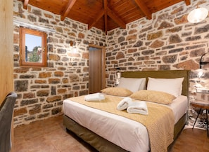 Un dormitorio con paredes de piedra, vigas de madera, una cama con ropa de cama estampada y una mesita con una lámpara.