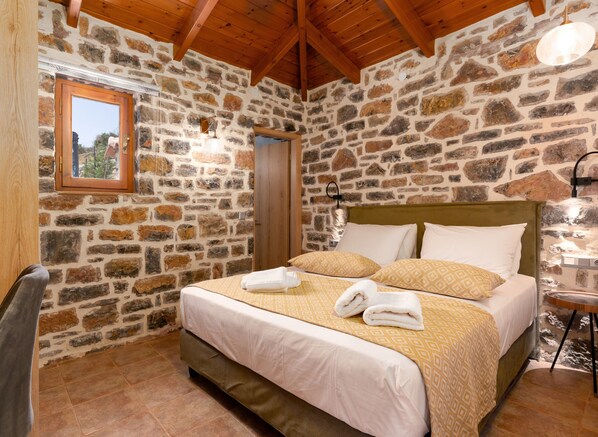 A bedroom with stone walls, wooden beams, a bed with patterned bedding, and a small table with a lamp.