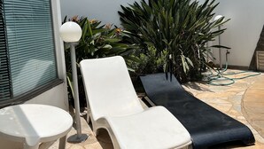 A patio with two white lounge chairs, a black lounge chair, and a white table. There are potted plants and a hose on the ground. - Espacial Motel (Aracatuba)