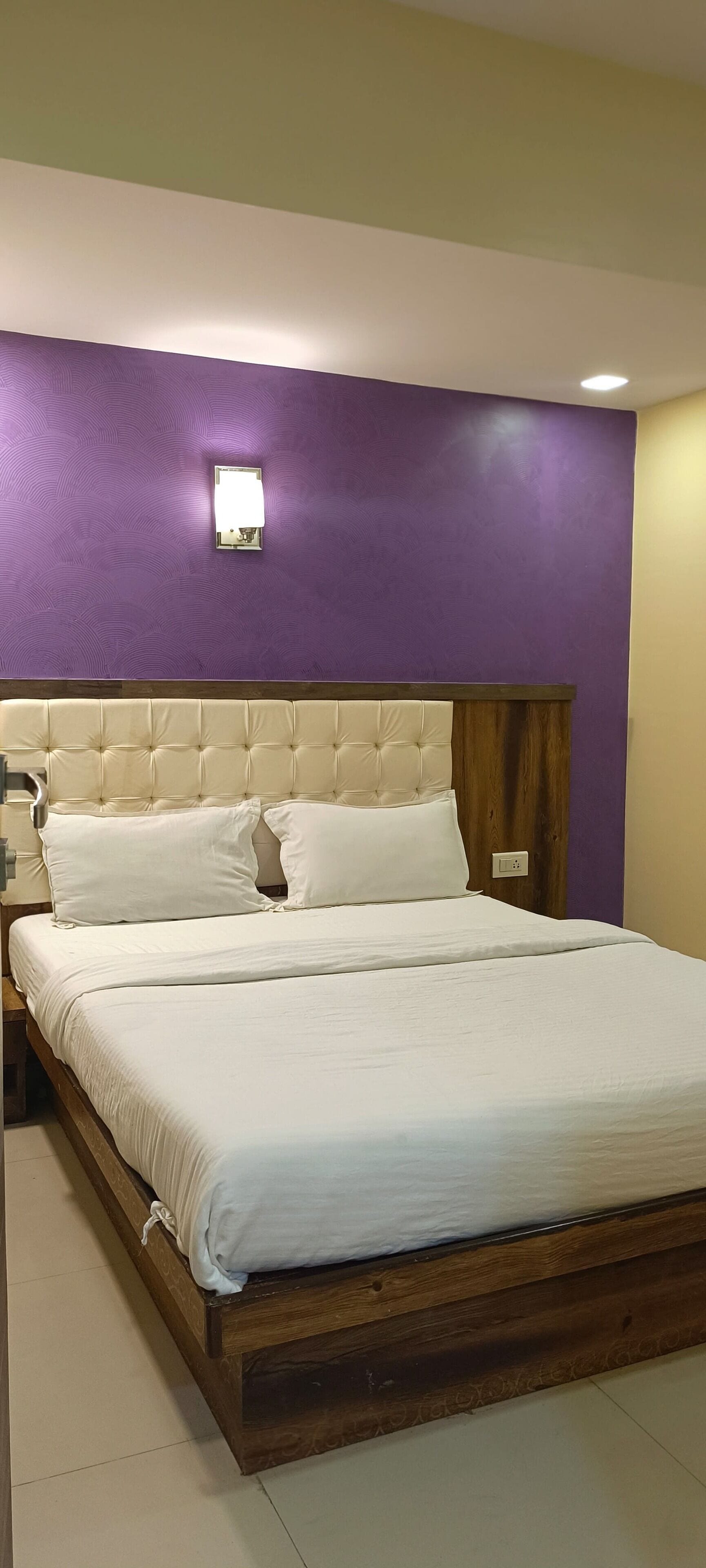 A neatly made bed with white linens, a wooden headboard, and a purple accent wall with a wall-mounted light fixture.