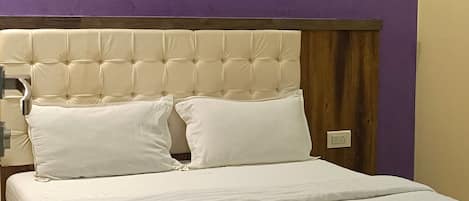 A neatly made bed with white linens, a wooden headboard, and a purple accent wall with a wall-mounted light fixture.