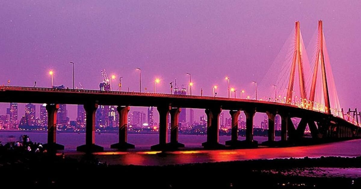 A bridge with multiple pillars and cables, spanning over a body of water with a city skyline in the background.