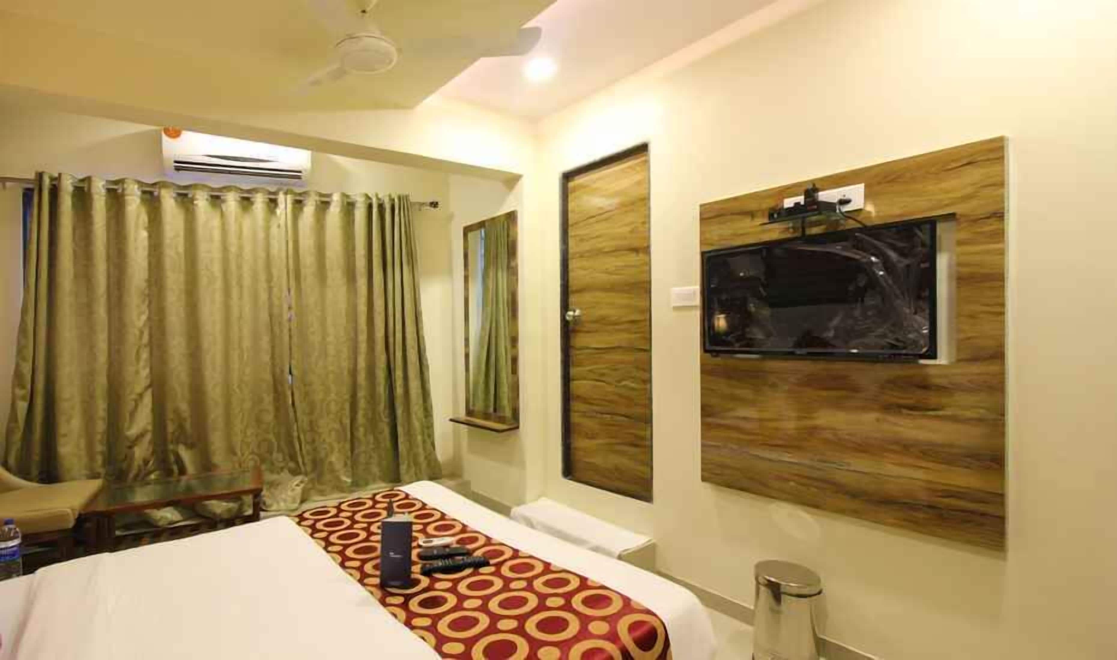 A hotel room with a bed, a TV mounted on the wall, a window with curtains, and an air conditioning unit.