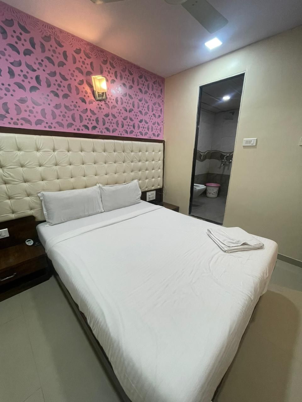 A hotel room with a large bed, a headboard, a nightstand, a wall-mounted light, and a bathroom door.