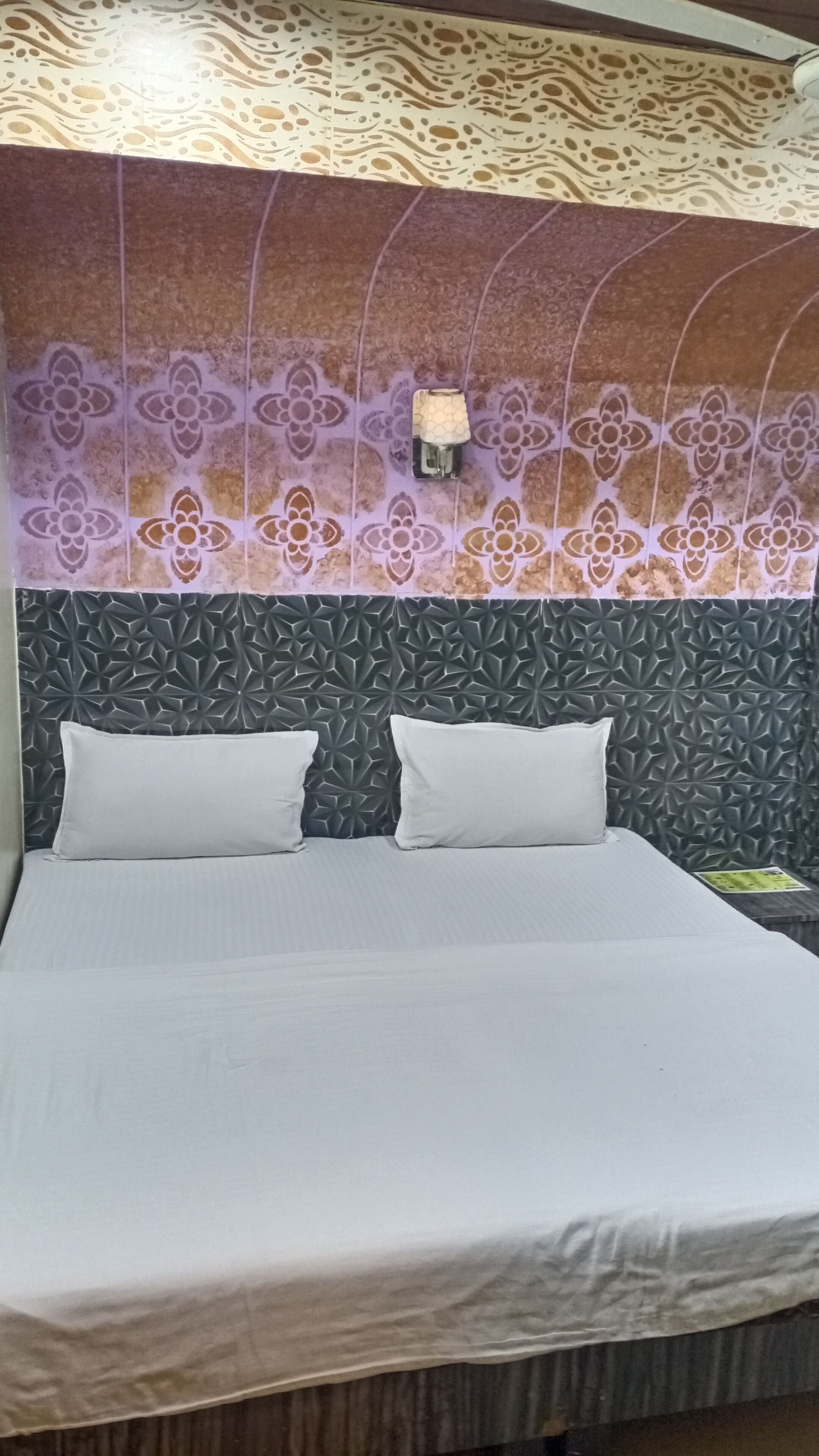 A bed with a white mattress and two pillows, a textured wall with a floral pattern, and a wall-mounted light fixture.