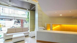 A modern reception area with a white reception desk, a seating area with white sofas, and a large window providing a view of the street outside. - Hotel Western Court  (Mumbai)