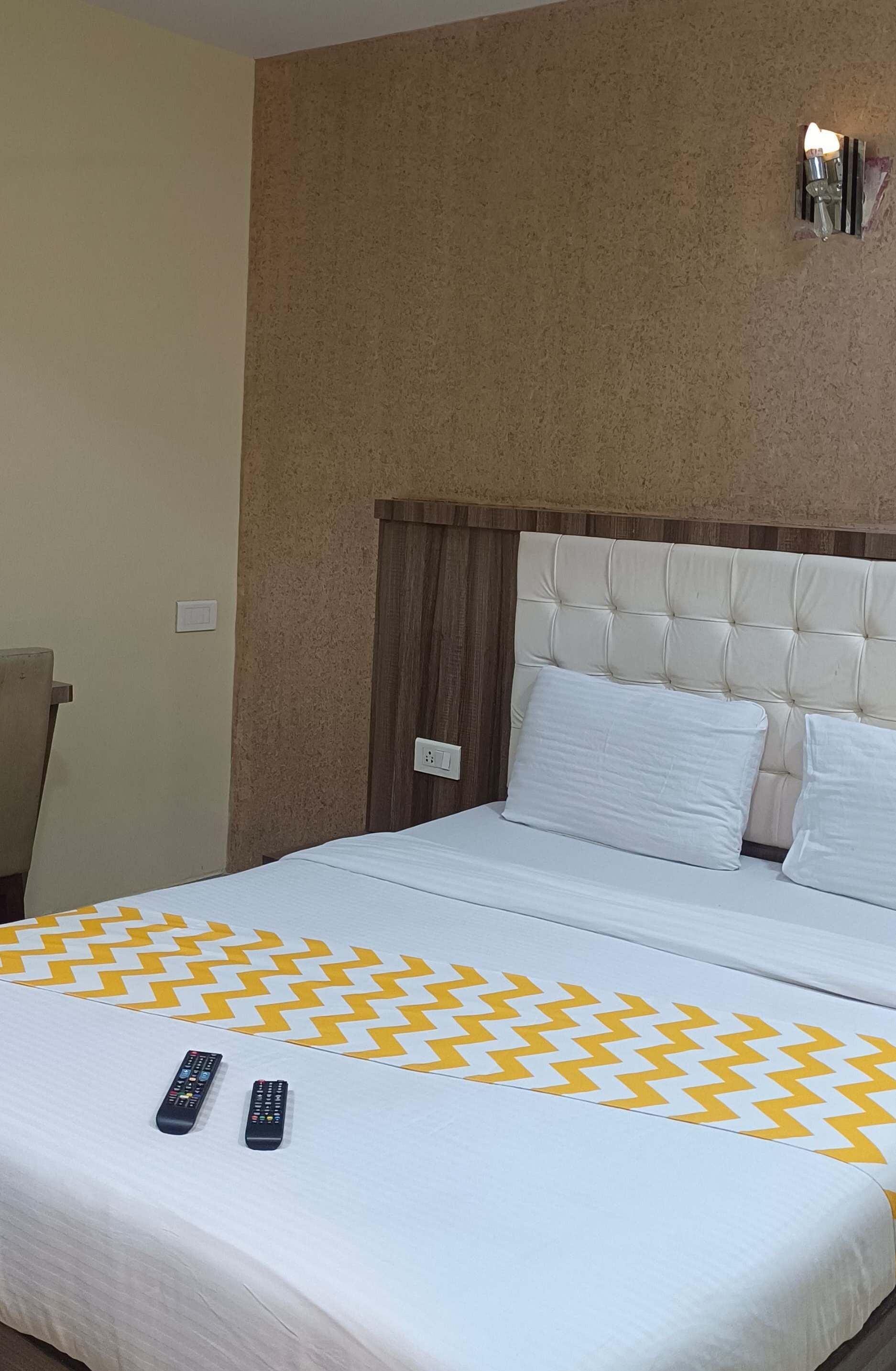 A hotel room with a bed, two remote controls, a wall-mounted light, and a chair.