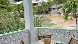 A round wooden table with four chairs, set on a balcony with a white railing and a view of a garden and buildings.