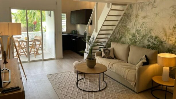 A modern living room with a sofa, a coffee table, and a TV. There is a staircase leading to another level and a dining area with chairs and a table.