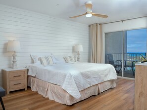 Une chambre avec un lit, des tables de chevet, un ventilateur de plafond, un balcon avec des chaises et une vue sur l’océan.