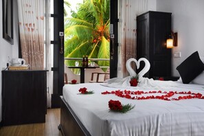 Una cama bien hecha con una toalla en forma de corazón y pétalos de flores rojas, una mesita con una tetera y botellas, y una vista de palmeras a través de una ventana.