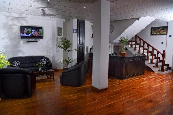 Un salón espacioso con un sofá negro, una mesa de centro y un televisor montado en la pared. Se ve una escalera con barandilla de madera que conduce a un nivel superior.
