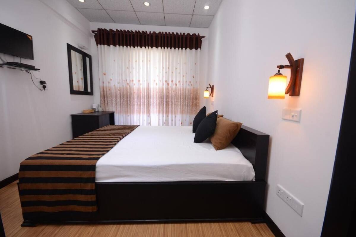 A bedroom with a large bed, a bedside table, a TV mounted on the wall, and a window with curtains.