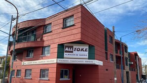 A red building with a sign for 'ALL TOKE ALMACÉN DE BARRIO' and a list of items like 'FIAMBRES - PANIFICADOS - BEBIDAS'. - Confortable departamento en Mendoza (Mendoza)