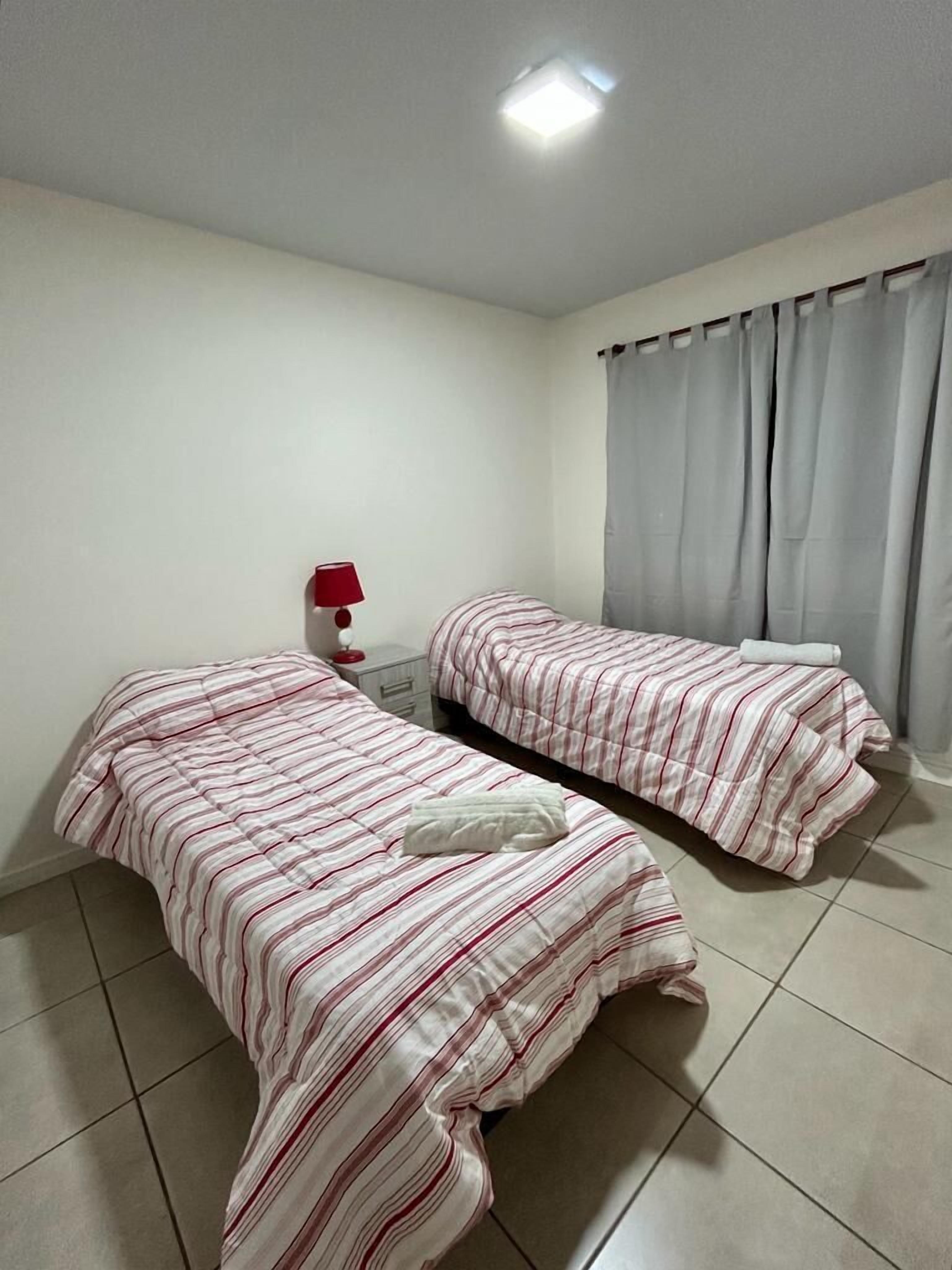 A room with two beds, a nightstand, a lamp, and a window with curtains.
