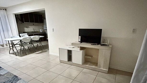 A living room with a TV on a stand, a dining table with chairs, and a kitchen area in the background. - Confortable departamento en Mendoza (Mendoza)