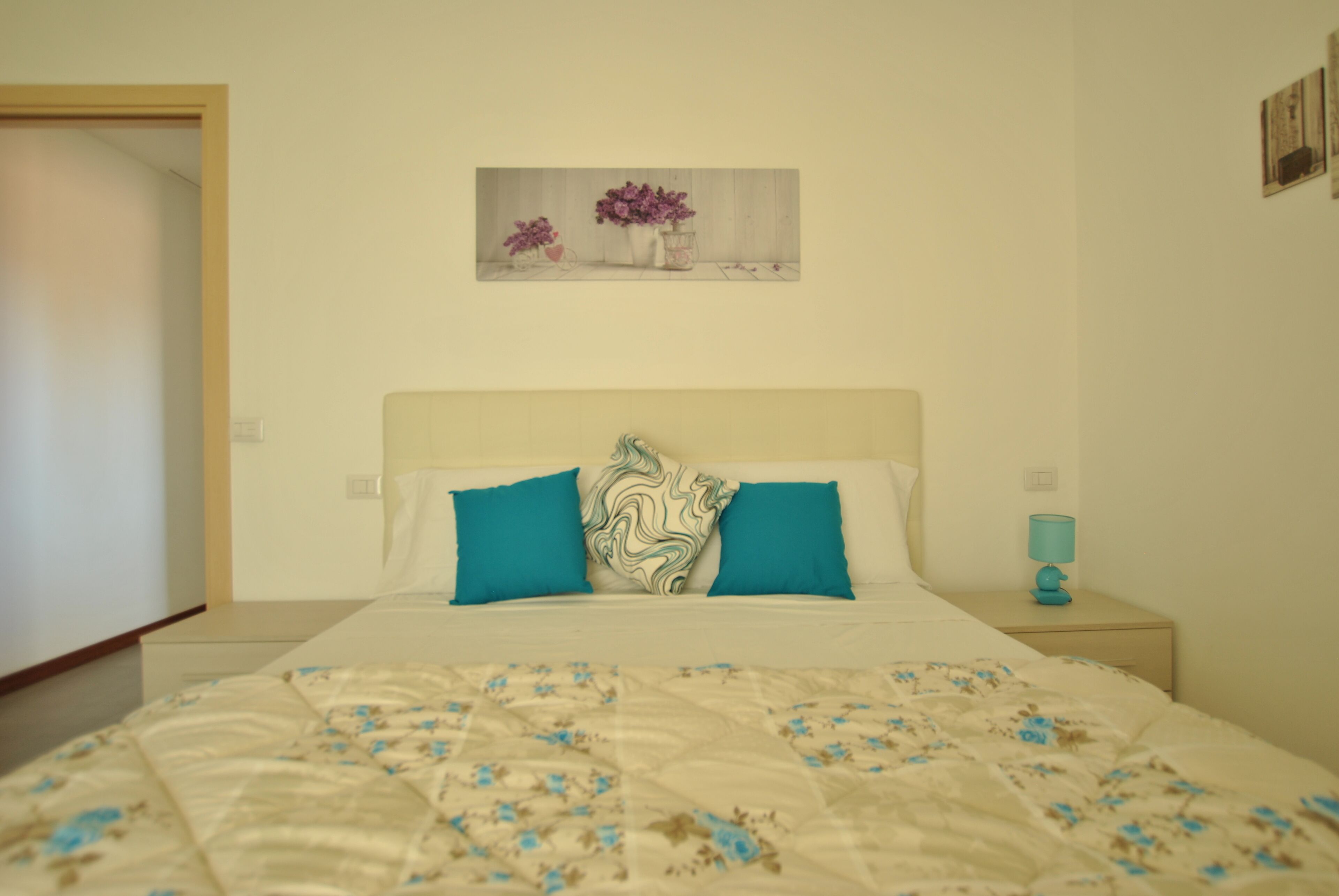 Een slaapkamer met een bed, twee kussens, een nachtkastje met een lamp en een aan de muur hangend schilderij.