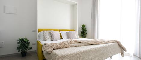 A bedroom with a bed, a bedside plant, and a window with curtains.