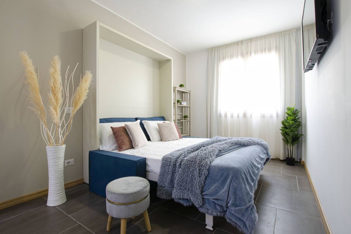 Un dormitorio con una cama, una mesita de noche, una planta y una estantería con objetos decorativos.