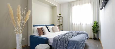A bedroom with a bed, a side table, a plant, and a shelf with decorative items.