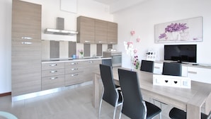 Una cocina moderna con electrodomésticos integrados, un comedor con una mesa larga y sillas, y un televisor de pantalla plana montado en la pared.