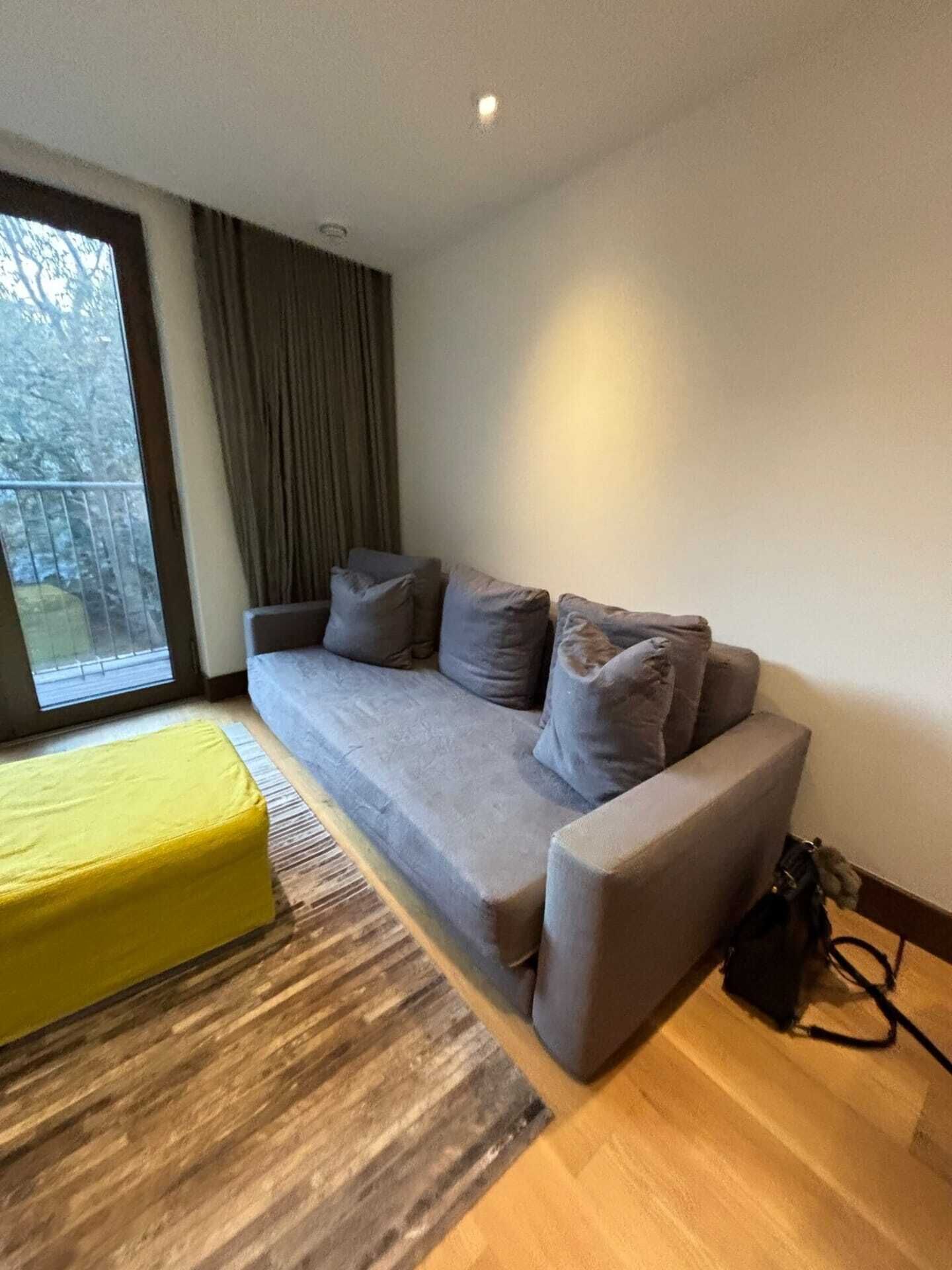 A modern living room with a grey sofa, a yellow ottoman, and a large window with curtains.