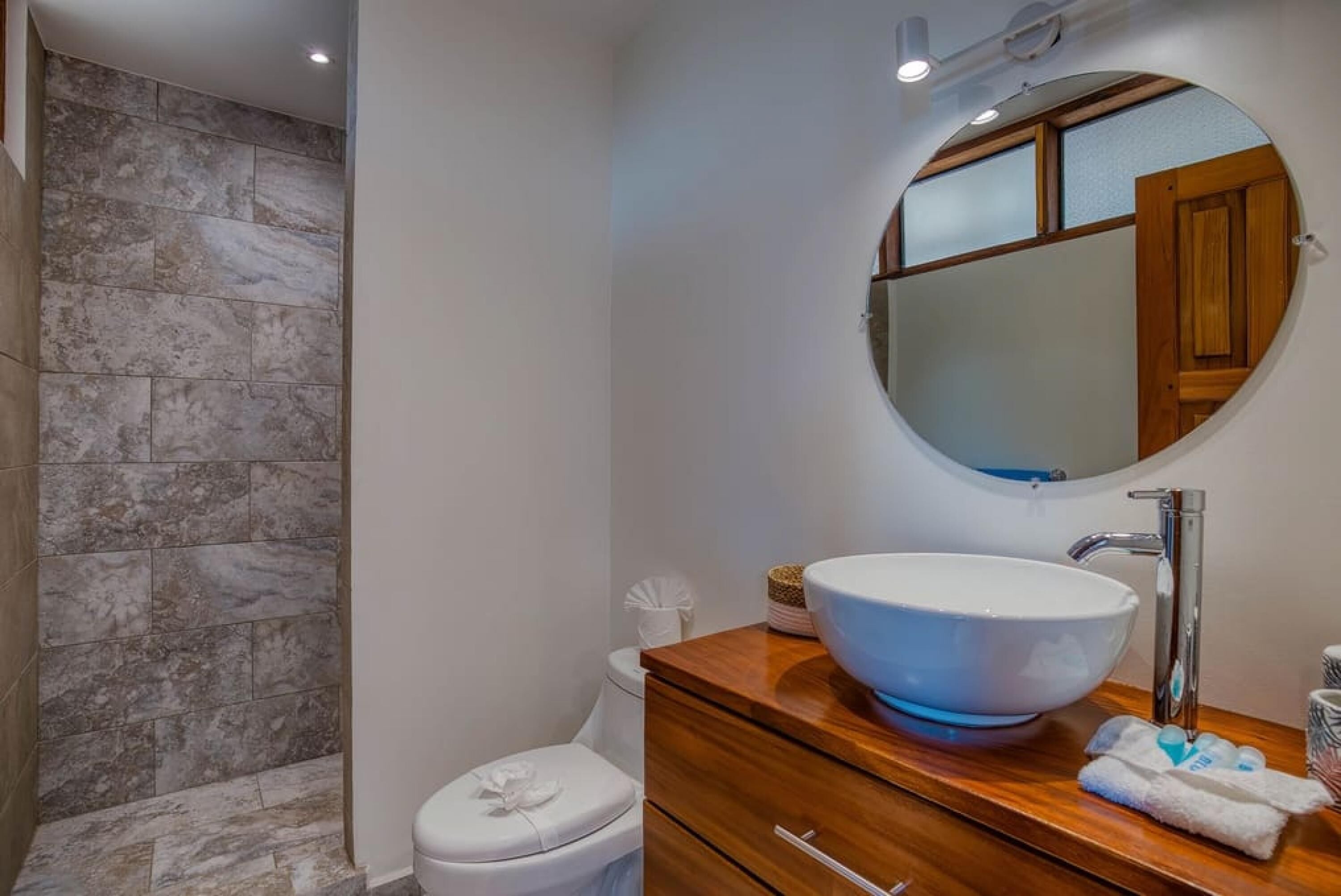 A bathroom with a white toilet, a wooden vanity with a large round mirror, and a vessel sink.