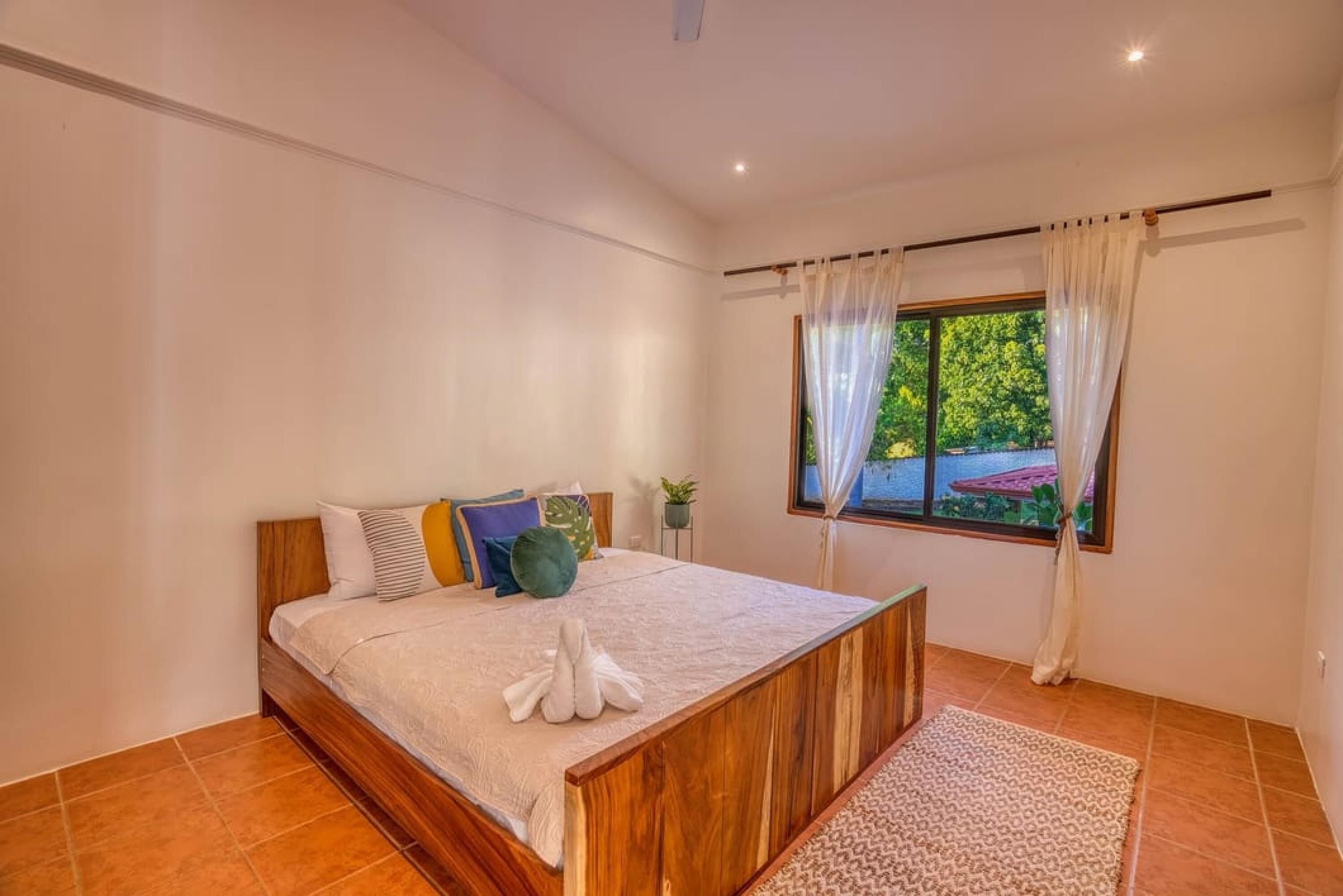 A bedroom with a wooden bed, a view of greenery outside, and a window with sheer curtains.