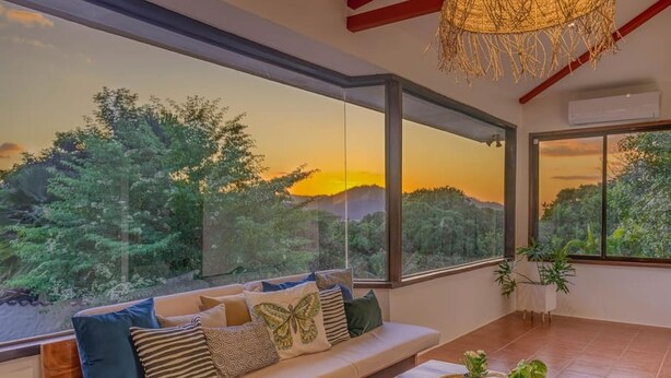 A modern living room with a large window offering a scenic view of trees and hills during sunset. - Few Steps to Beach Private Pool and Jacuzzi (Sardinal)