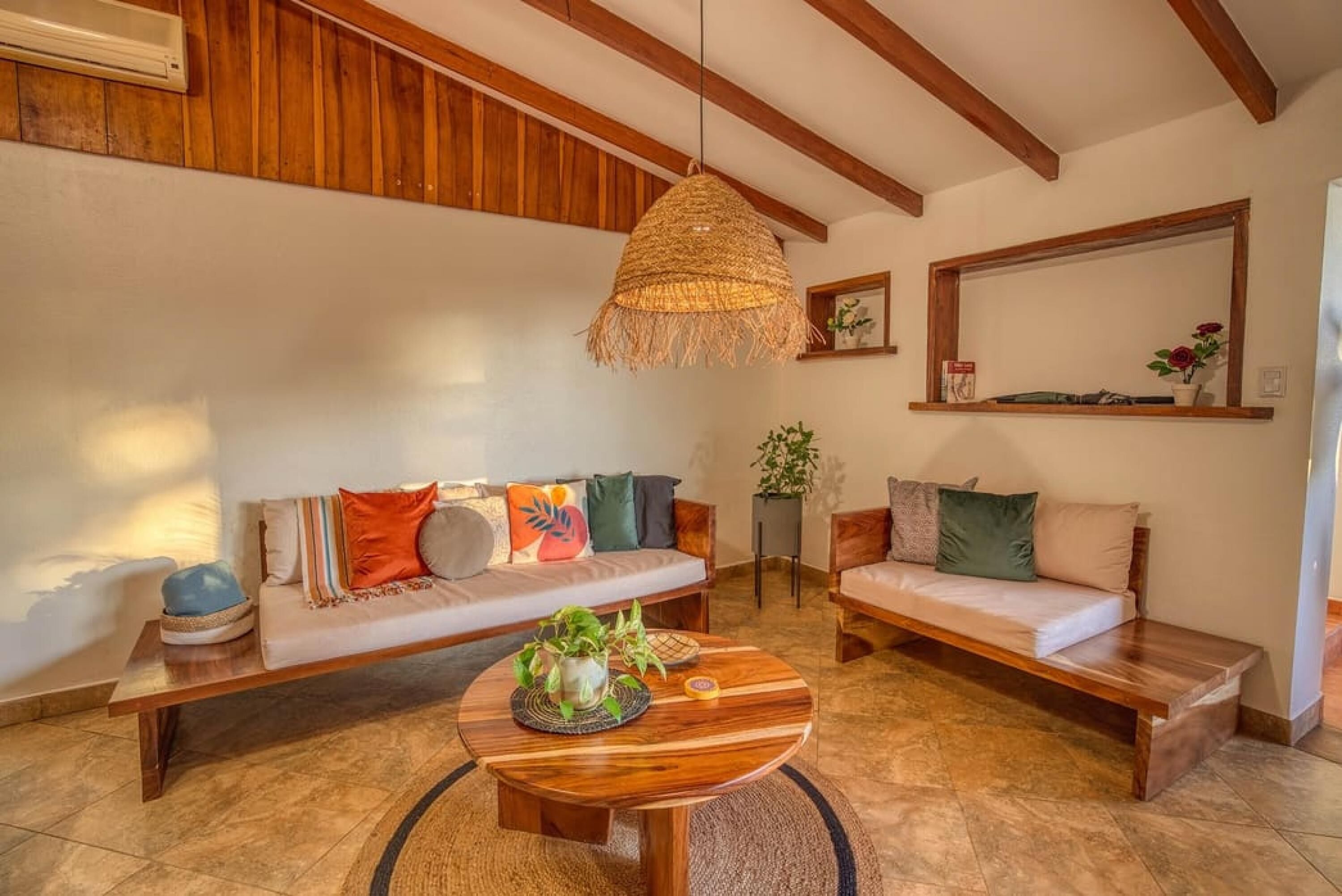 Un salon chaleureux avec un plafond en bois, un luminaire suspendu en fibres tressées, un canapé agrémenté de coussins colorés, un banc et une table basse ronde ornée de plantes.