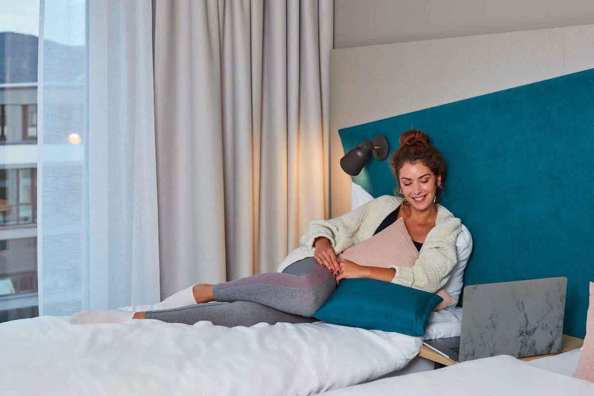 Une personne assise sur un lit, avec un ordinateur portable, des oreillers et une couverture.