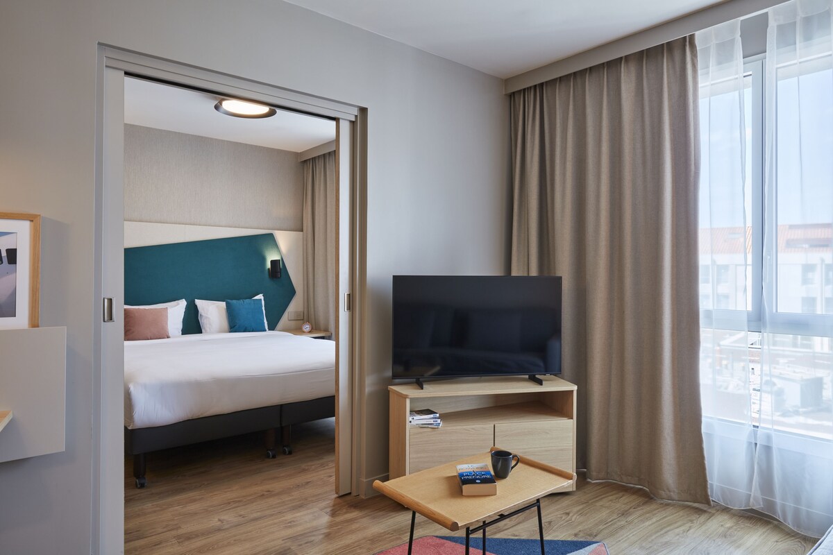 A hotel room with a bed, a TV on a wooden stand, a small table with a book and a mug, and a window with curtains.