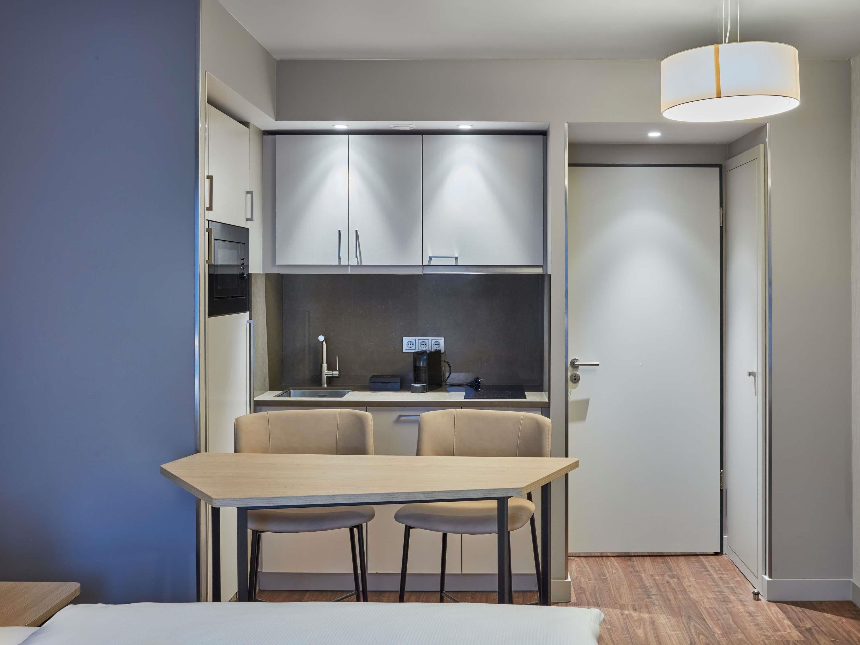 A compact kitchenette with a microwave, sink, and coffee maker, a dining table with two chairs, and a door leading to another room.