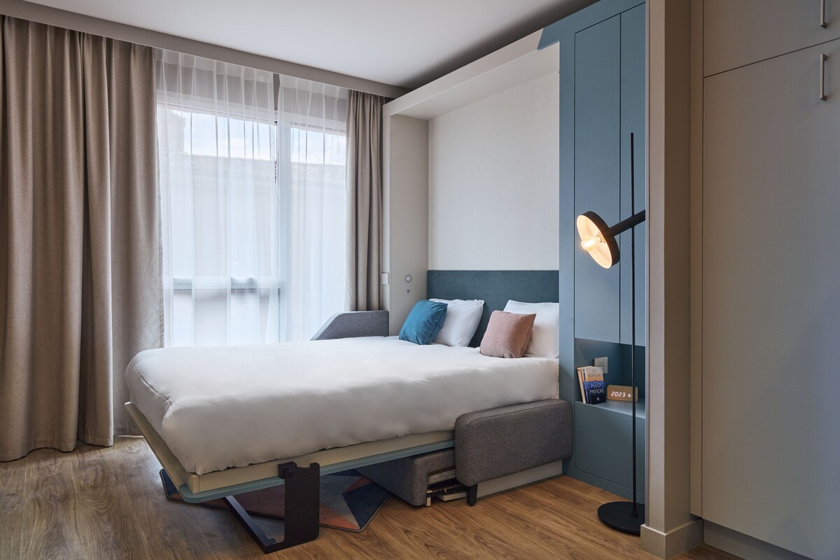 A modern hotel room with a bed, a desk, and a wardrobe.