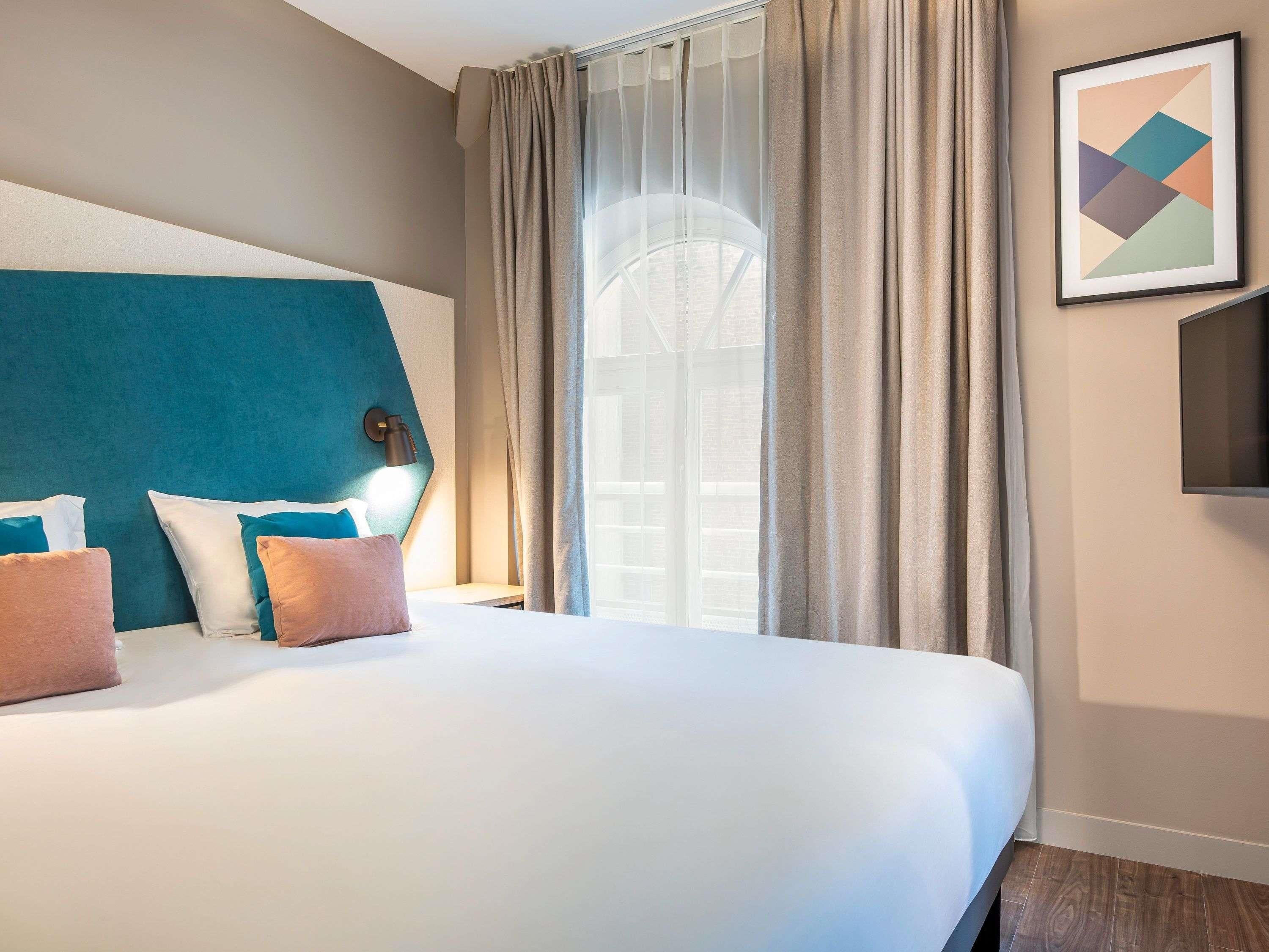 Une chambre d’hôtel avec un grand lit, une tête de lit, un téléviseur fixé au mur et une fenêtre avec des rideaux.