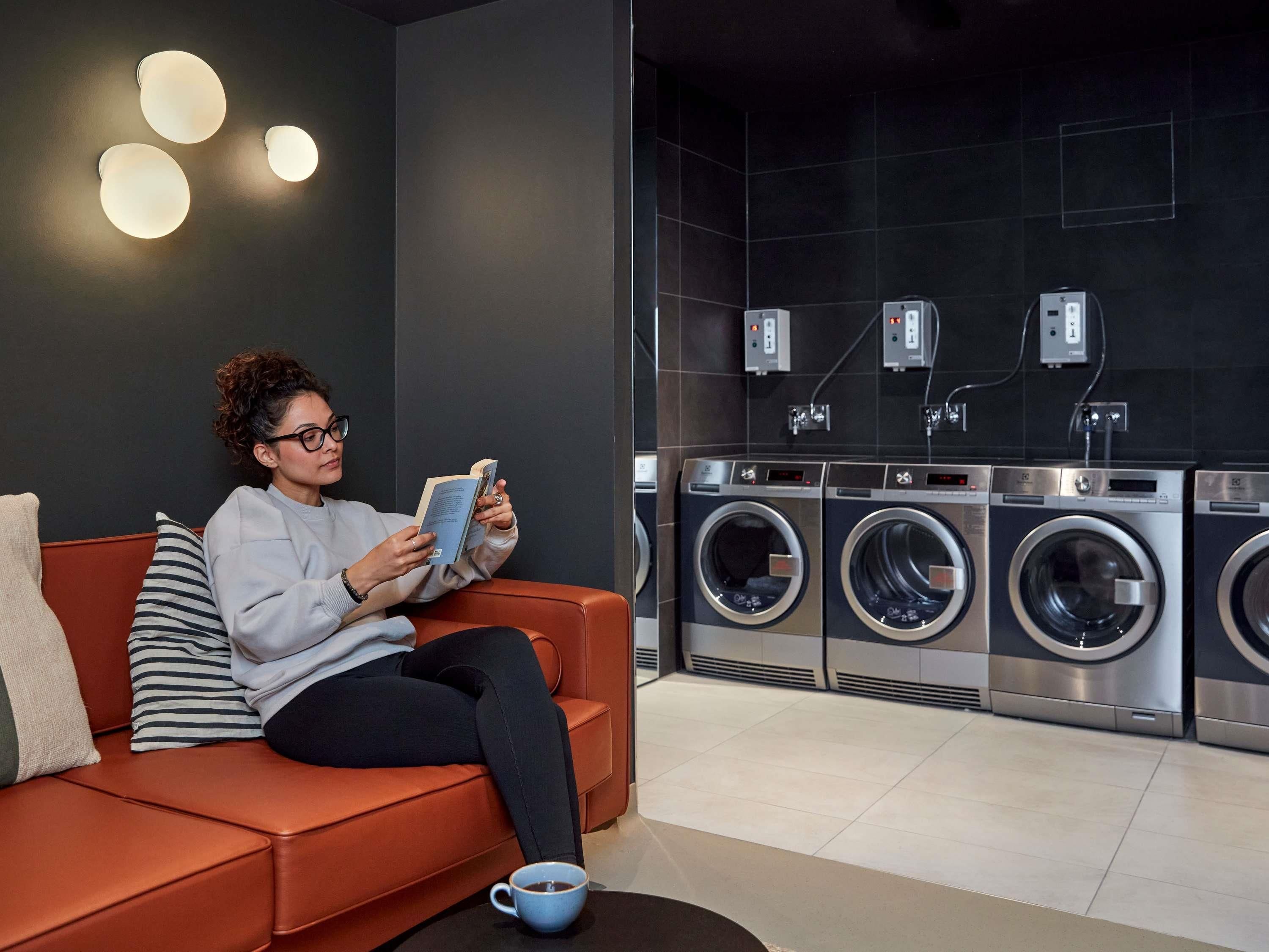 A person sitting on a sofa reading a book in a modern room with a washing machine and a coffee cup on the floor.