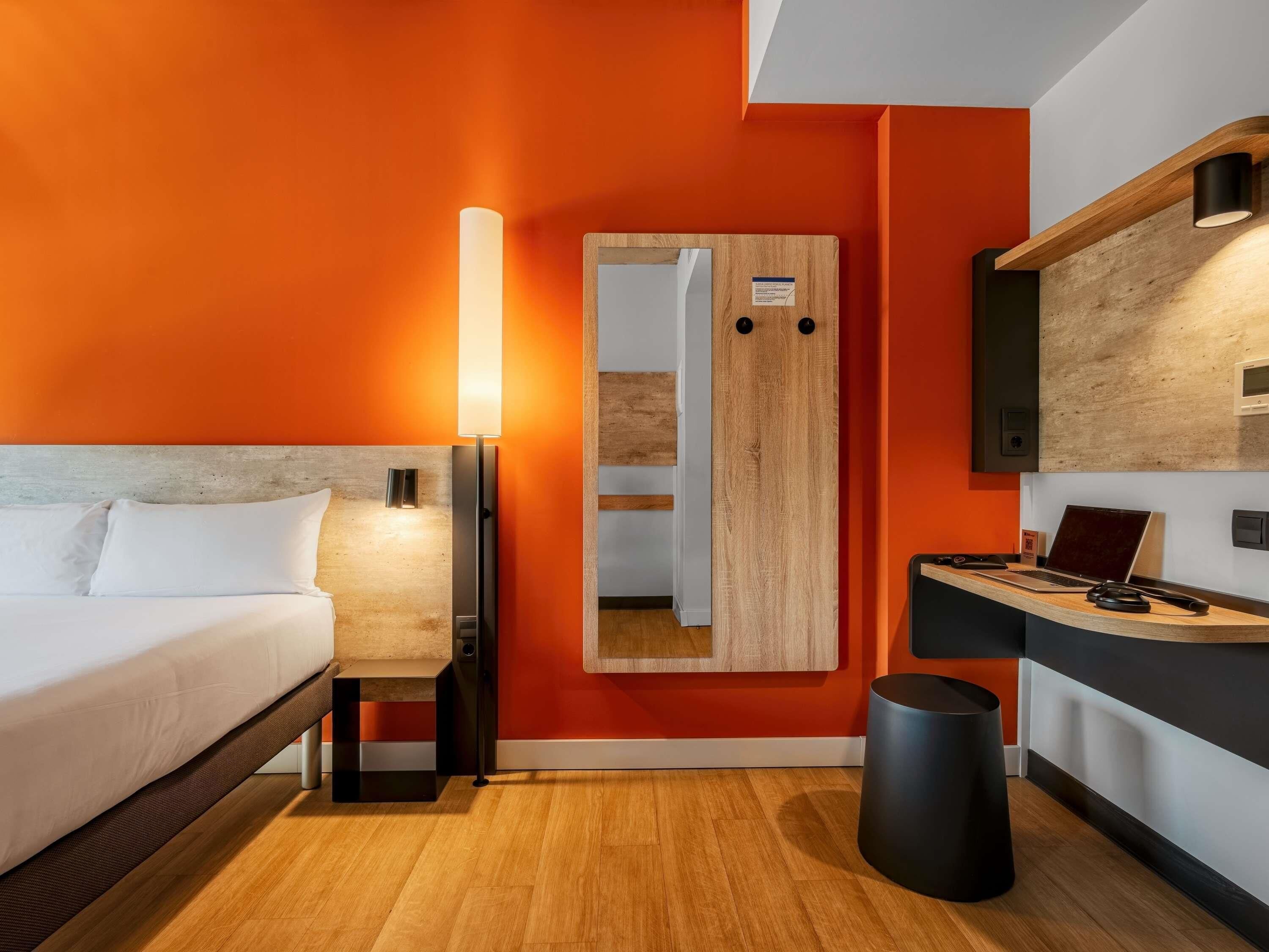 A modern hotel room with a bed, a desk with a laptop, a mirror, and a trash bin.