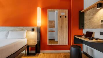 Une chambre d’hôtel moderne équipée d’un lit, d’un bureau avec un ordinateur portable, d’un miroir et d’une poubelle.