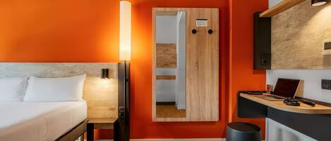 Una habitación de hotel moderna con una cama, un escritorio con una computadora portátil, un espejo y un bote de basura.