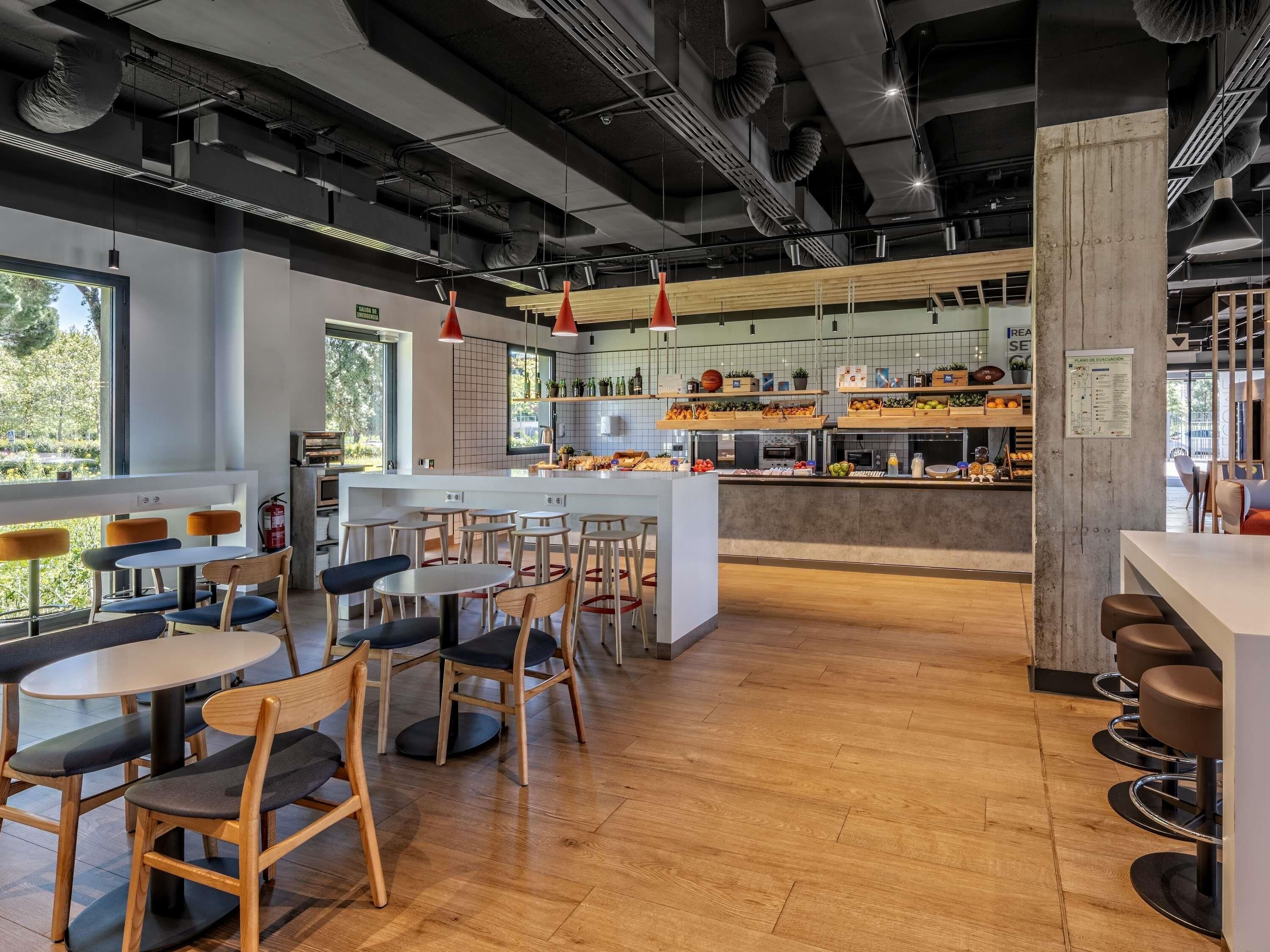 Interior de una cafetería moderna con mesas y sillas de madera, una barra central con estantes llenos de tentempiés y vistas al exterior con vegetación.