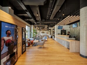 Un área de recepción moderna con un mostrador de madera, zona de estar con sillas y mesas, y un cartel grande en la pared.