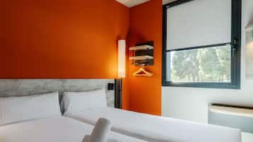 A hotel room with a bed, a window with a view of trees, a wall-mounted light, and a wooden shelf.