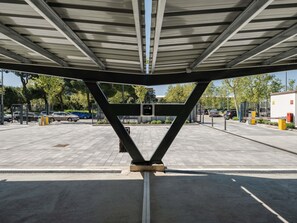 A parking garage entrance with a metal truss structure, a pedestrian walkway, and a gated area with vehicles. - Ibis Budget Madrid Aeropuerto (Madrid)