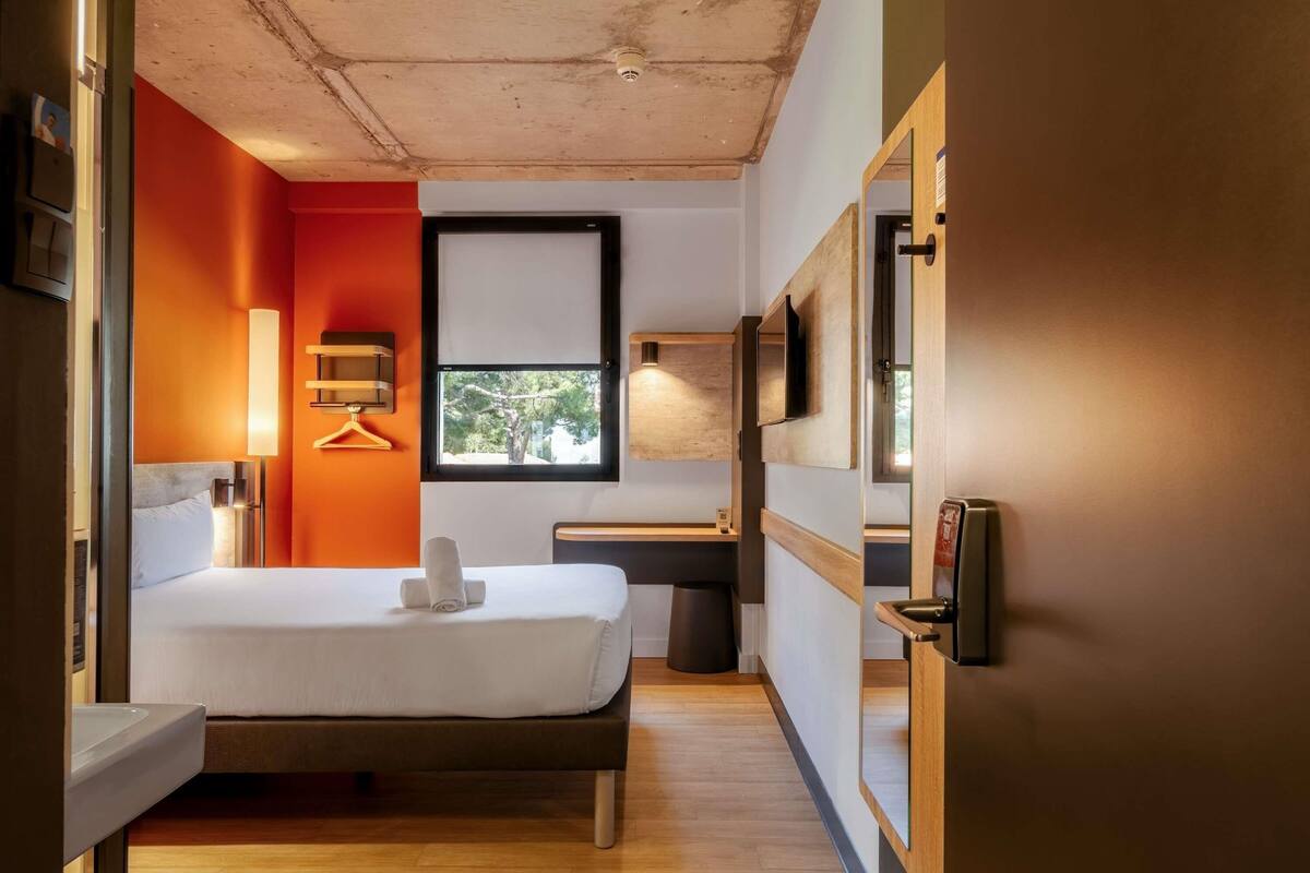 A modern hotel room with a bed, bedside table, lamp, and a view of greenery outside.