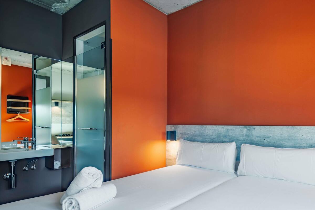 A modern hotel room with a large bed, a bathroom with a mirror, and a towel on the bed.