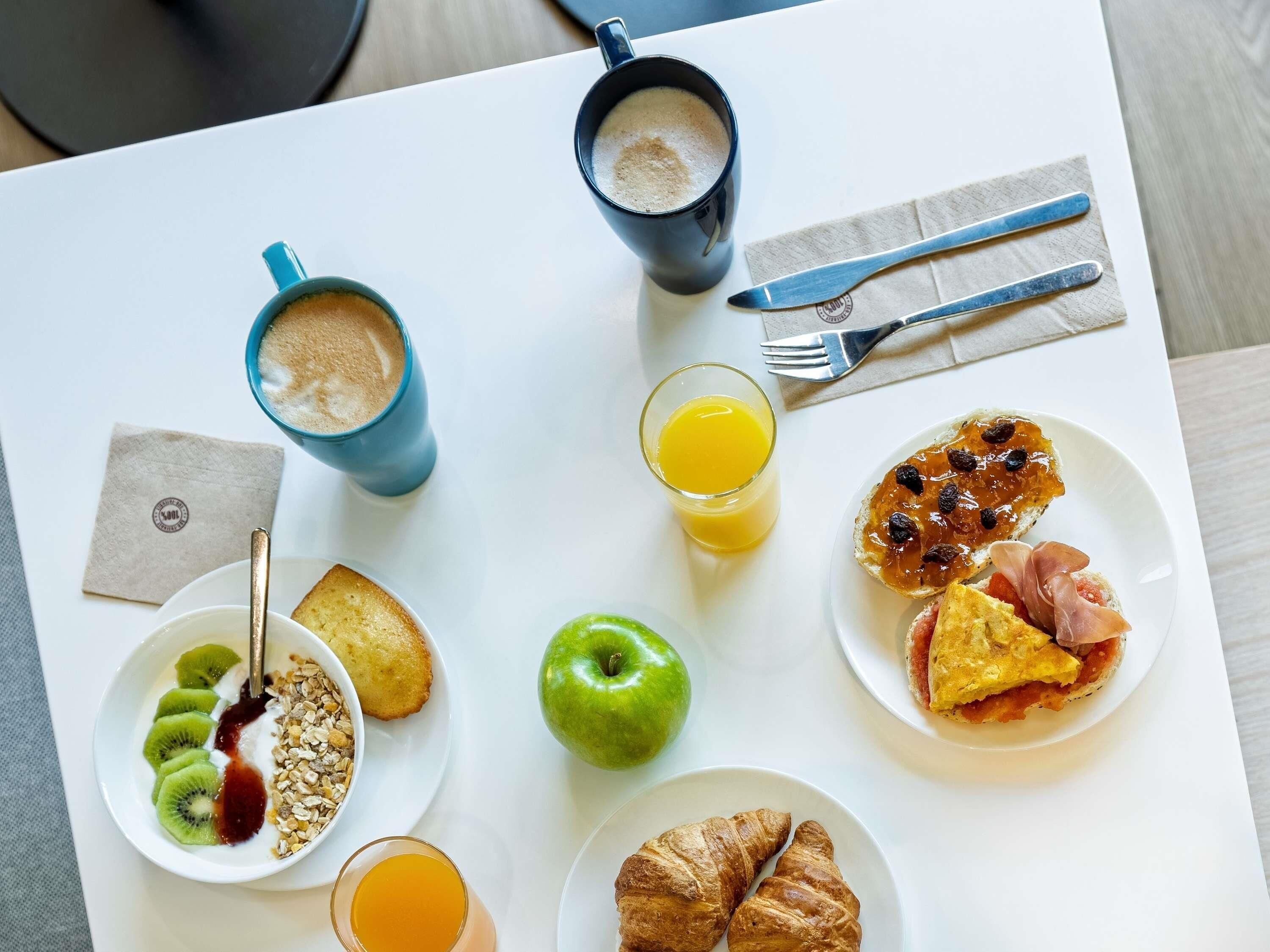Uma mesa de pequeno-almoço com vários alimentos e bebidas, incluindo um croissant, uma maçã verde e um copo de sumo de laranja.