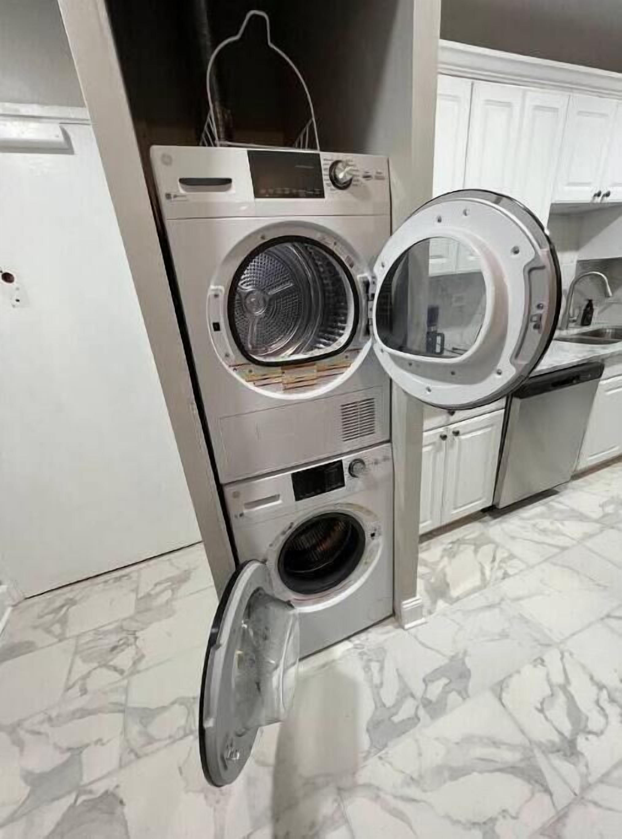 Un appareil combiné lave-linge et sèche-linge compact, en acier inoxydable, à chargement frontal, installé dans une cuisine avec des meubles blancs et un sol en marbre.